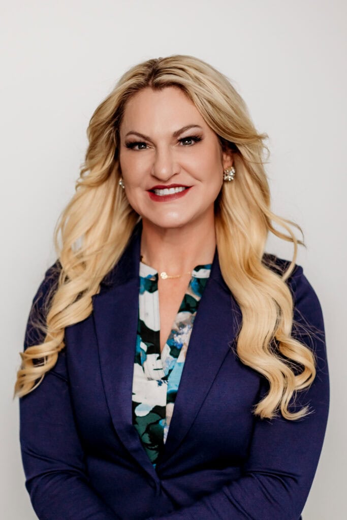 Professional portrait of a smiling woman with long blonde hair, wearing a blue blazer and a floral top.