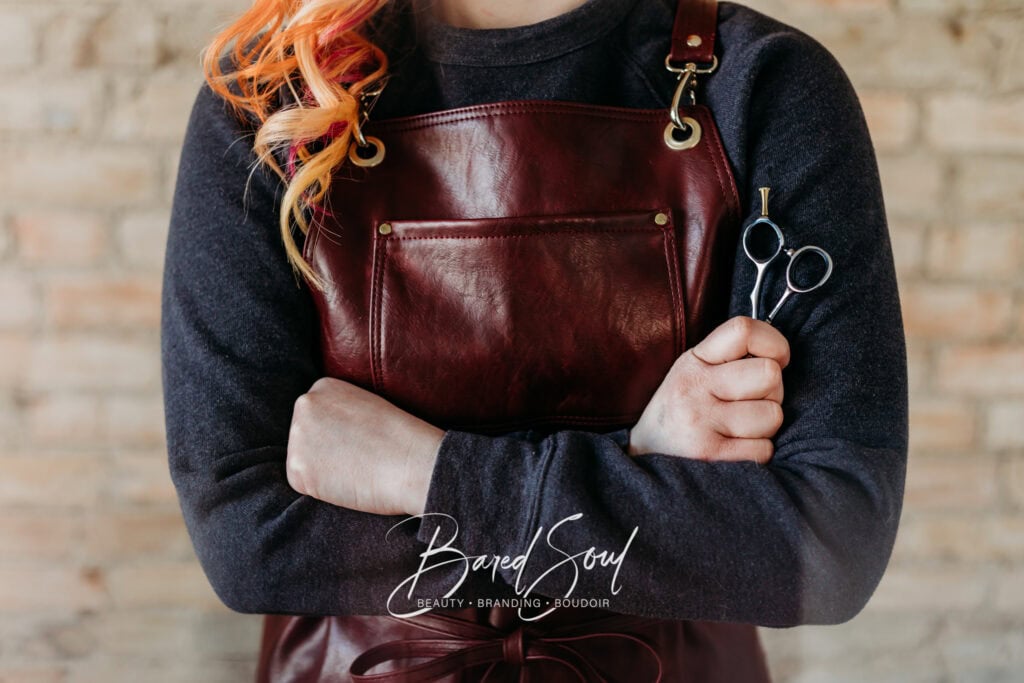 A person holding a brown leather bag and a pair of scissors, wearing a dark sweater, standing against a brick wall.