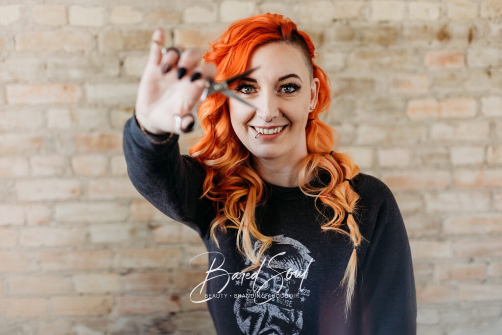 A woman with vibrant red hair smiling at the camera, holding a small object between her fingers, standing against a brick wall.