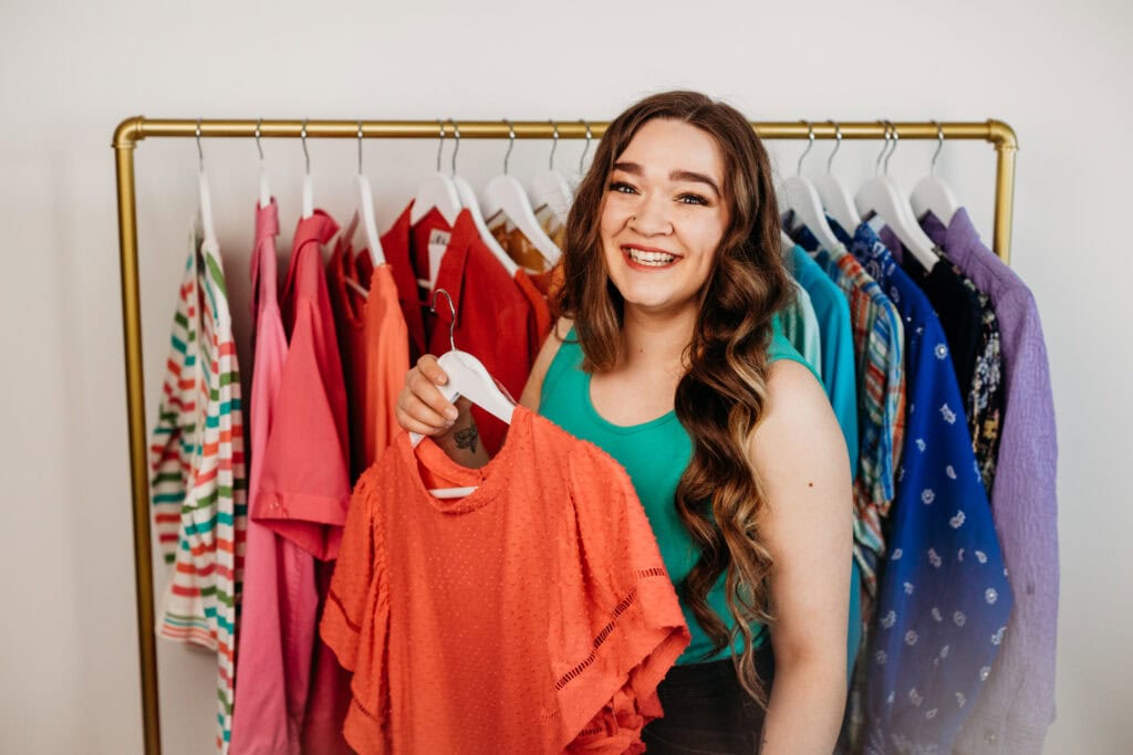 A woman with long brown hair smiling and holding a red shirt while standing next to a colorful clothing rack.