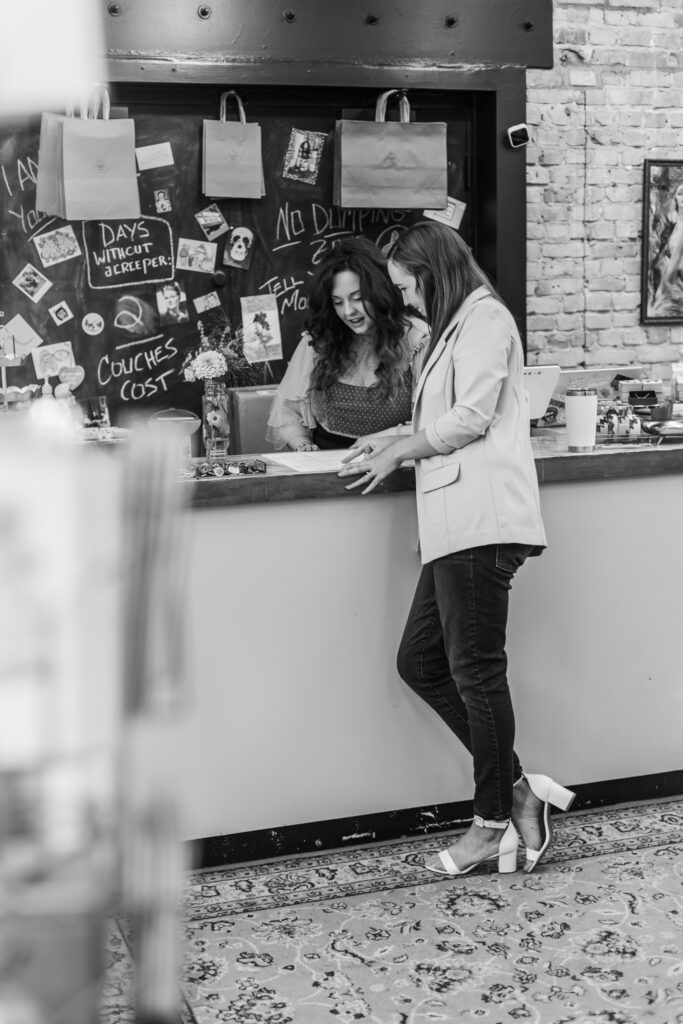 Two women talking and laughing at a cafe counter, surrounded by hanging paper bags and a chalkboard with doodles and writings.