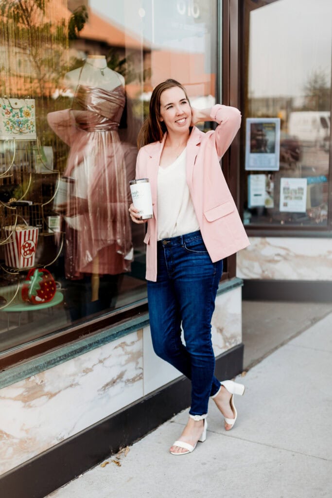A woman in a pink blazer and jeans smiles while holding a coffee cup, standing outside a boutique with mannequins in the window.