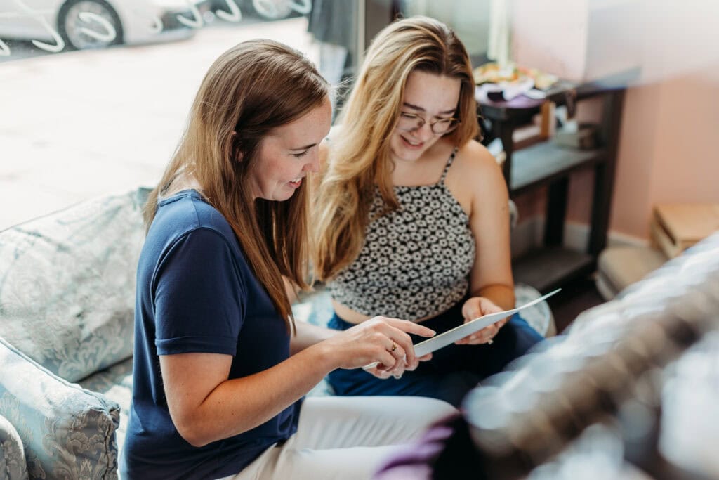 Two women sit on a sofa while looking at a tablet and smiling in a casual indoor setting.
