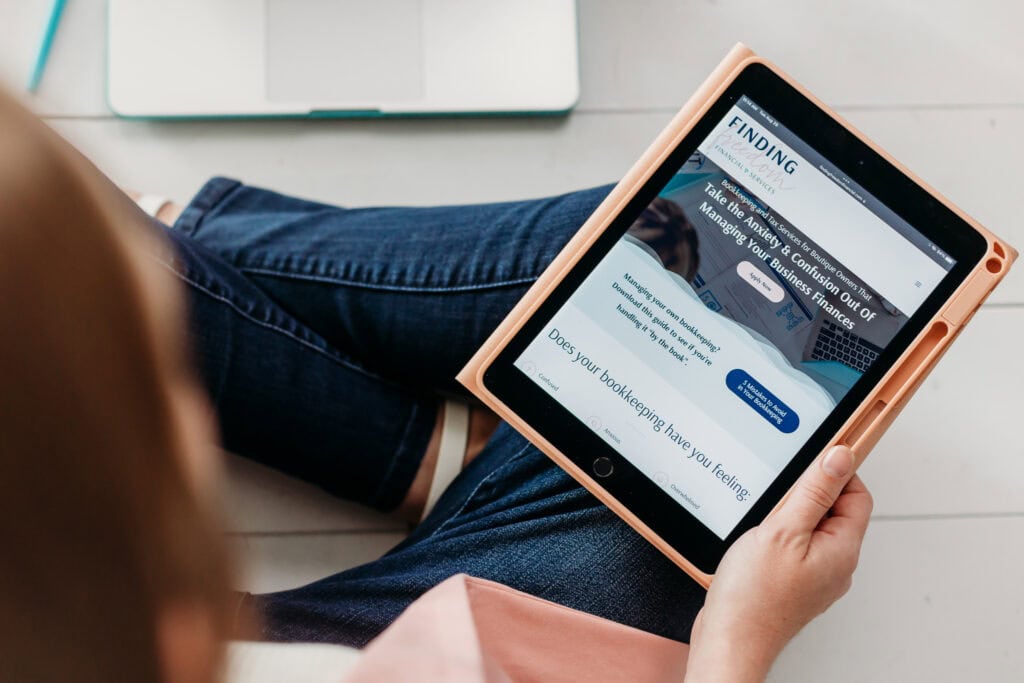 Person sitting on the floor with a tablet displaying a webpage about booking travel, with laptops visible in the background.