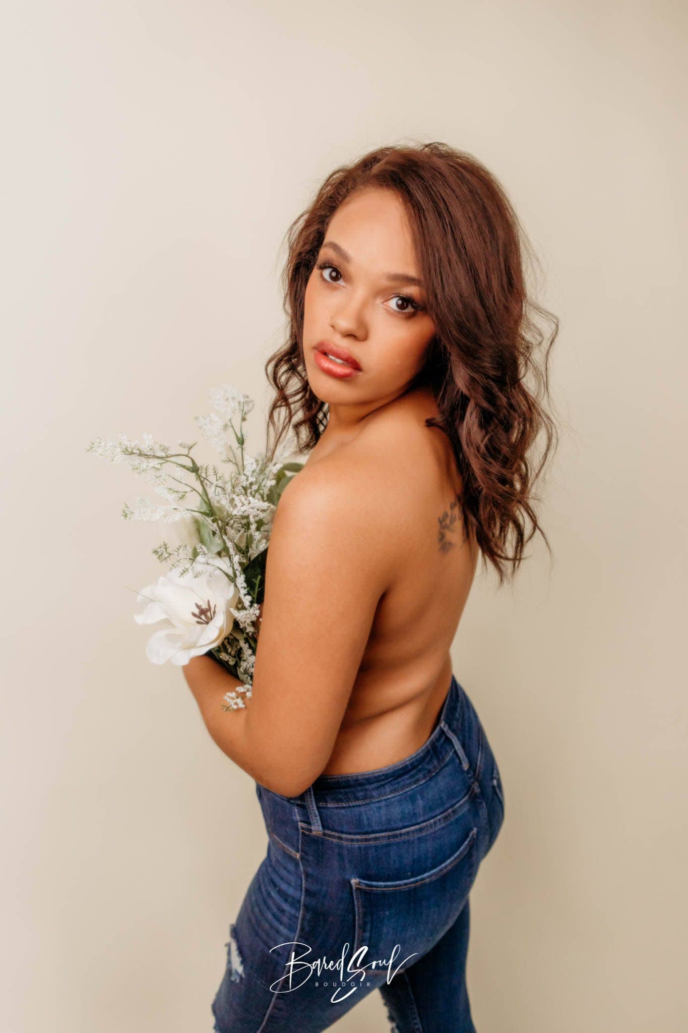 Woman with a subtle smile, holding white flowers, standing against a beige background, wearing blue jeans and looking over her shoulder.